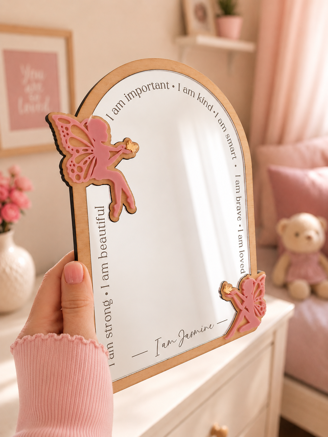 Pink fairy mirror with motivational phrases held by a hand in a room with decor elements.