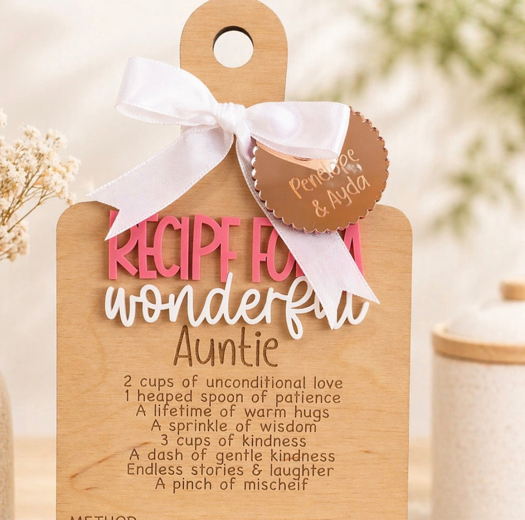 A wooden recipe card gift tag for an auntie, decorated with a white ribbon and a round brown paper tag that reads 'Pembje & Ayda'. The card features a humorous recipe for 'wonderful Auntie' written in pink and white lettering on a light wood background.