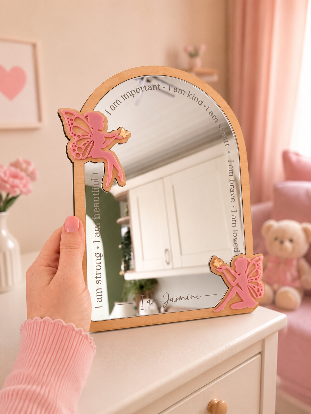 Decorative mirror with fairy design held by a hand in a room with pink decor.