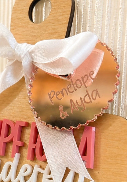 A close-up of a personalized wooden hanger with a white satin bow and a scalloped rose-gold tag that reads 'Penelope & Ayda'. The hanger is decorated with pink and white wooden letters spelling 'HPEFA dera' on a light wood background.