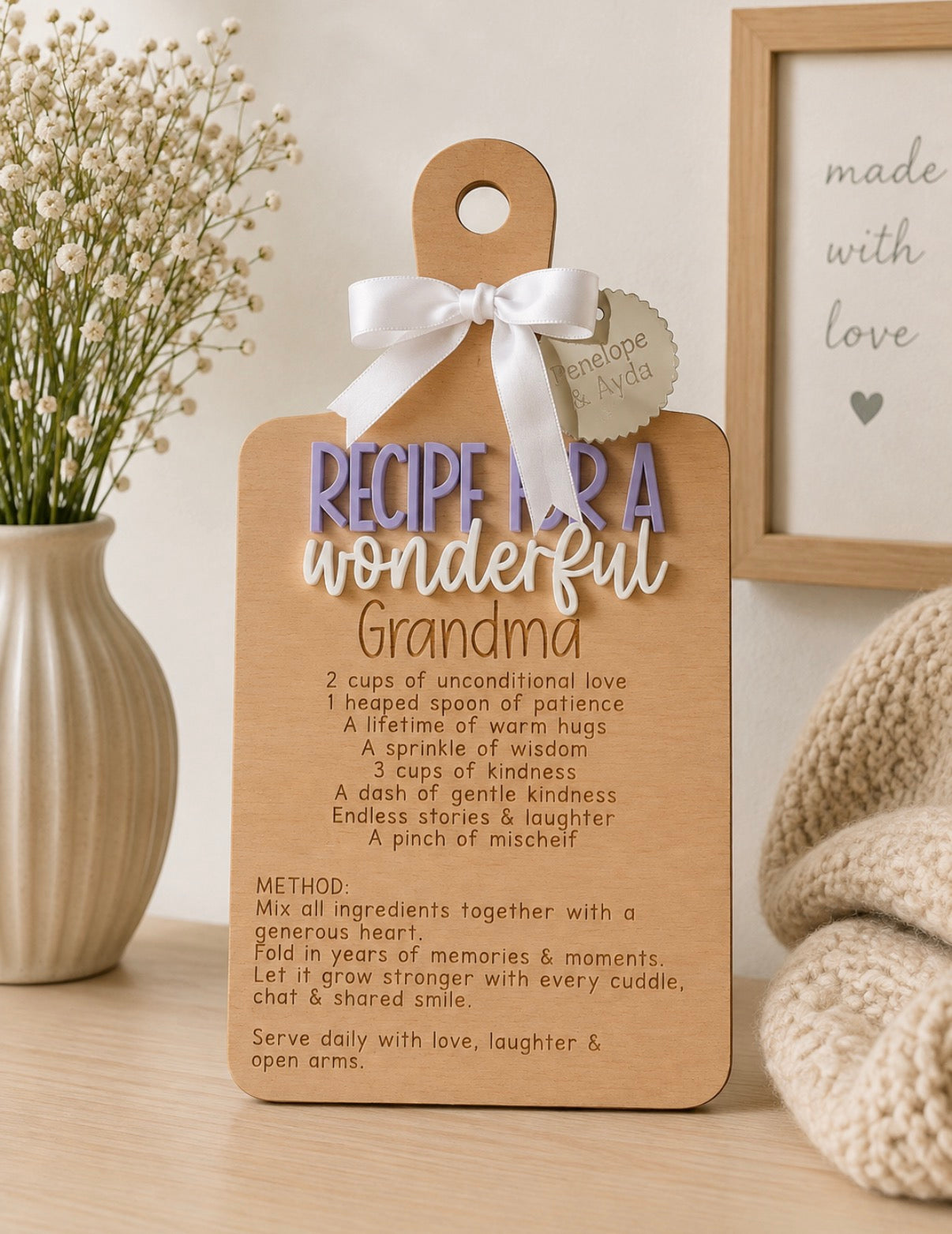 A wooden gift tag plaque with a white ribbon bow, printed with a recipe titled 'Recipe for a wonderful Grandma' and displayed on a tabletop with flowers and a framed print in the background.